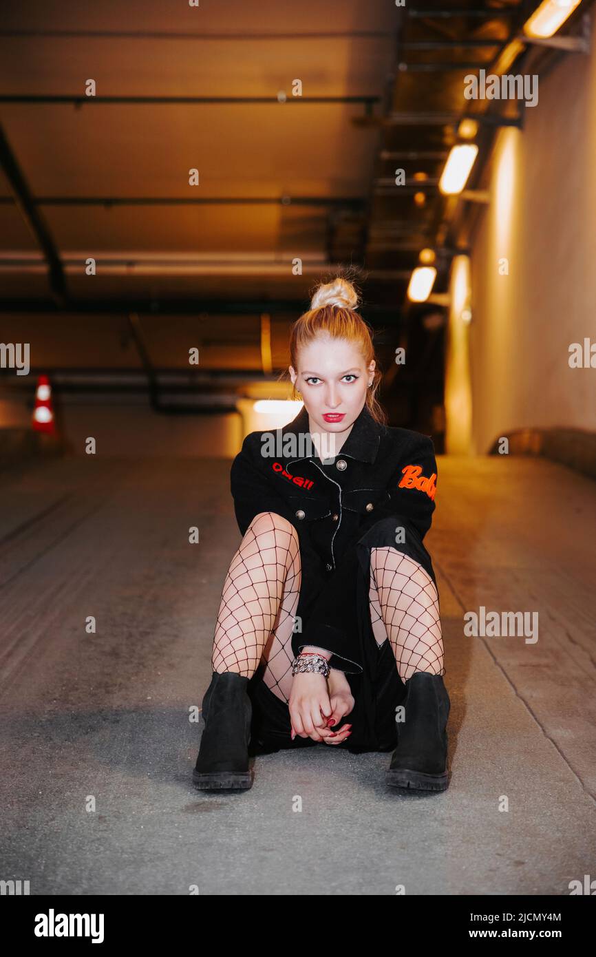 Bella ragazza bionda con capelli bun, indossando un cappotto nero, calze rete, si siede sottoterra sul pavimento di un garage parcheggio Foto Stock