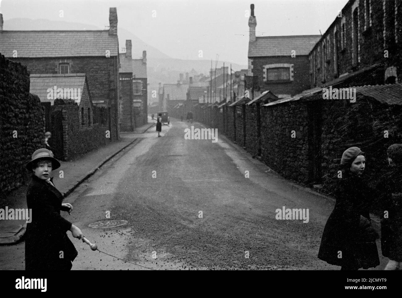 1950s, storico, una vista su una strada laterale umida nel villaggio gallese di coalmining Trehafod, Galles del Sud, con un po 'di scuola di bambini, uno tenendo la maniglia di una corda di salto, giocando all'esterno. Foto Stock