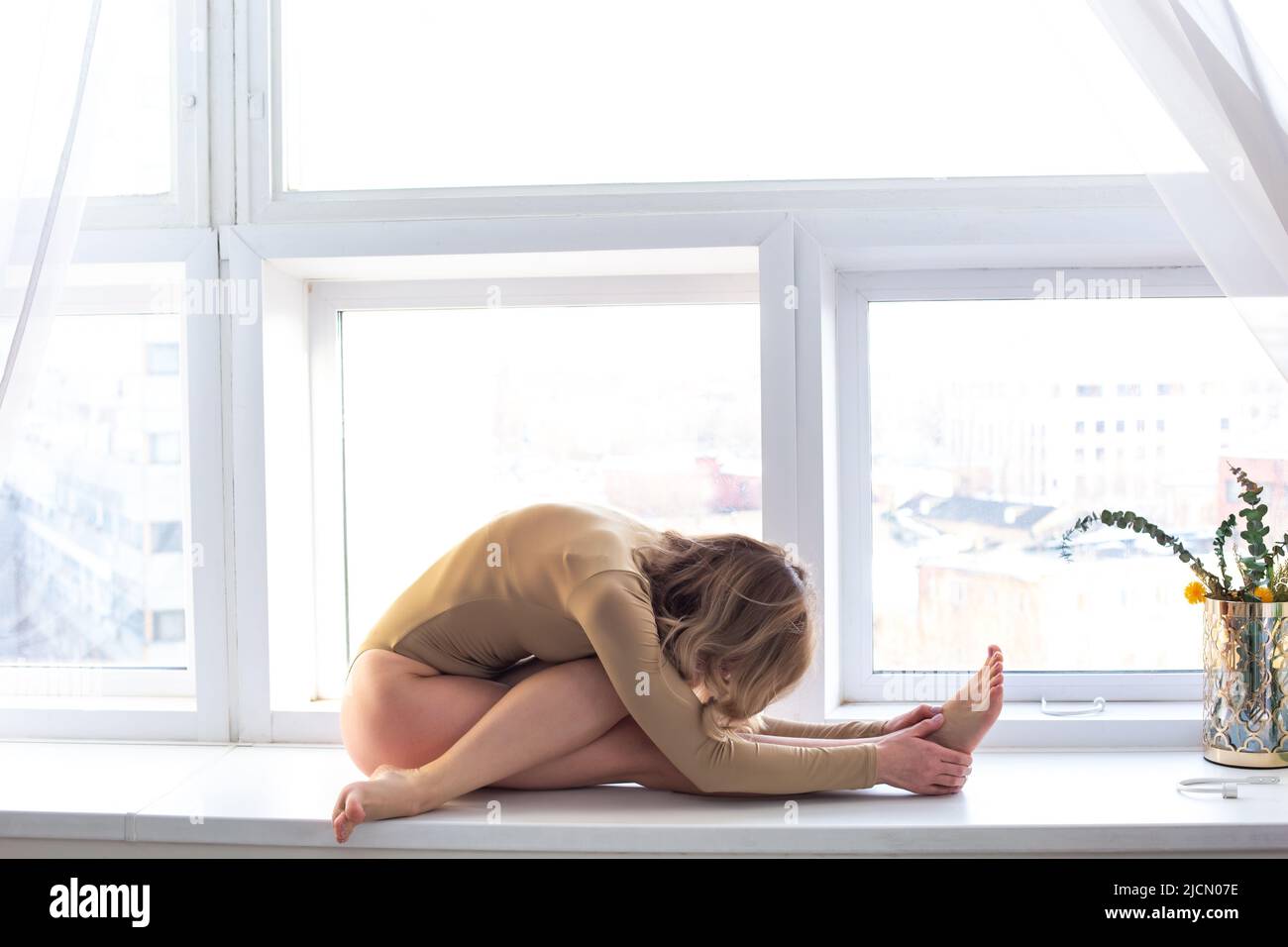 Una bella donna snella in un costume da bagno beige, si siede in una posa yoga vicino ad una grande finestra bianca Foto Stock