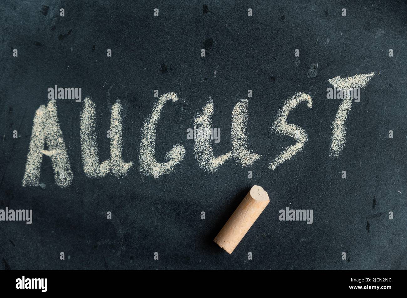 Agosto. Nome dell'ultimo mese estivo scritto in gesso giallo su lavagna nera. Testo scritto a mano. Un pezzo di gesso colorato si trova accanto ad esso. Foto Stock