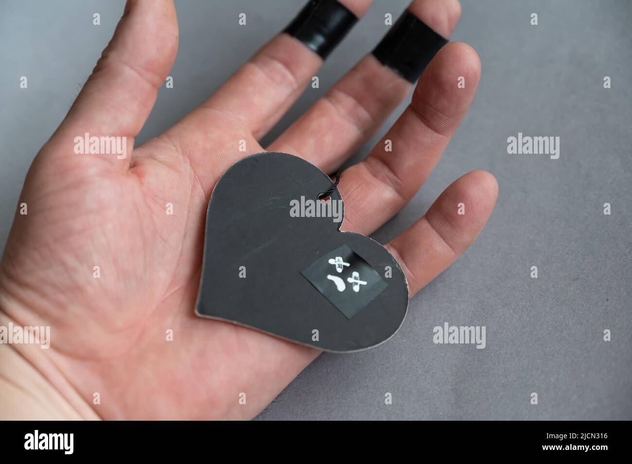 Un cuore nero si trova nel palmo di un uomo. Una figura di carta a forma di cuore con un volto tristemente disegnato. La mano di un uomo di mezza età. Amore non riacclamato, perdita di una lov Foto Stock