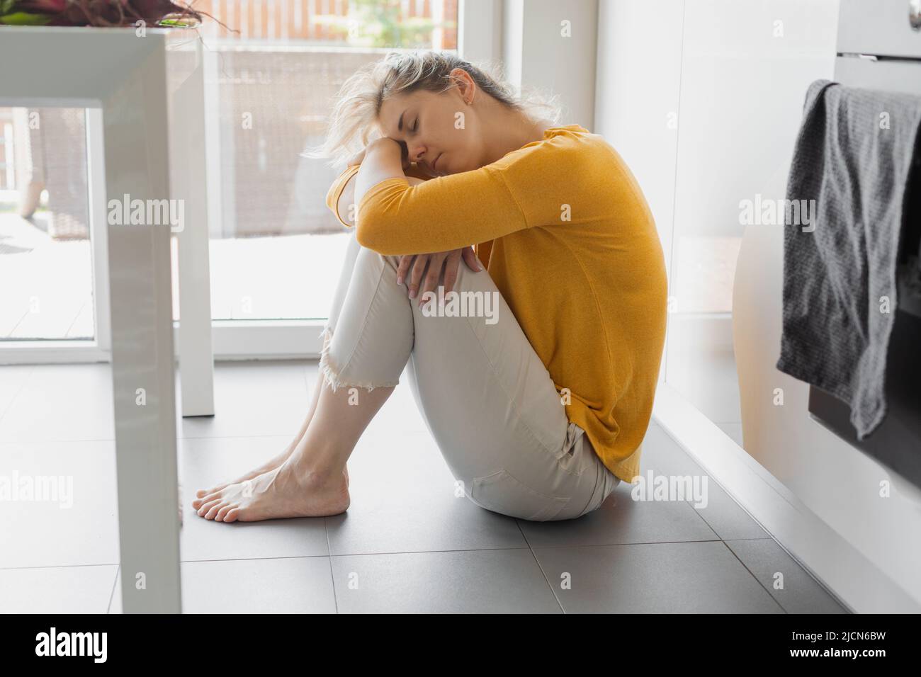 Depressi 30 anni donna seduta sul piano della cucina con ginocchia abbracciate. Solitudine, tristezza. Salute mentale, abuso di droga e concetto di problema sociale. Foto Stock