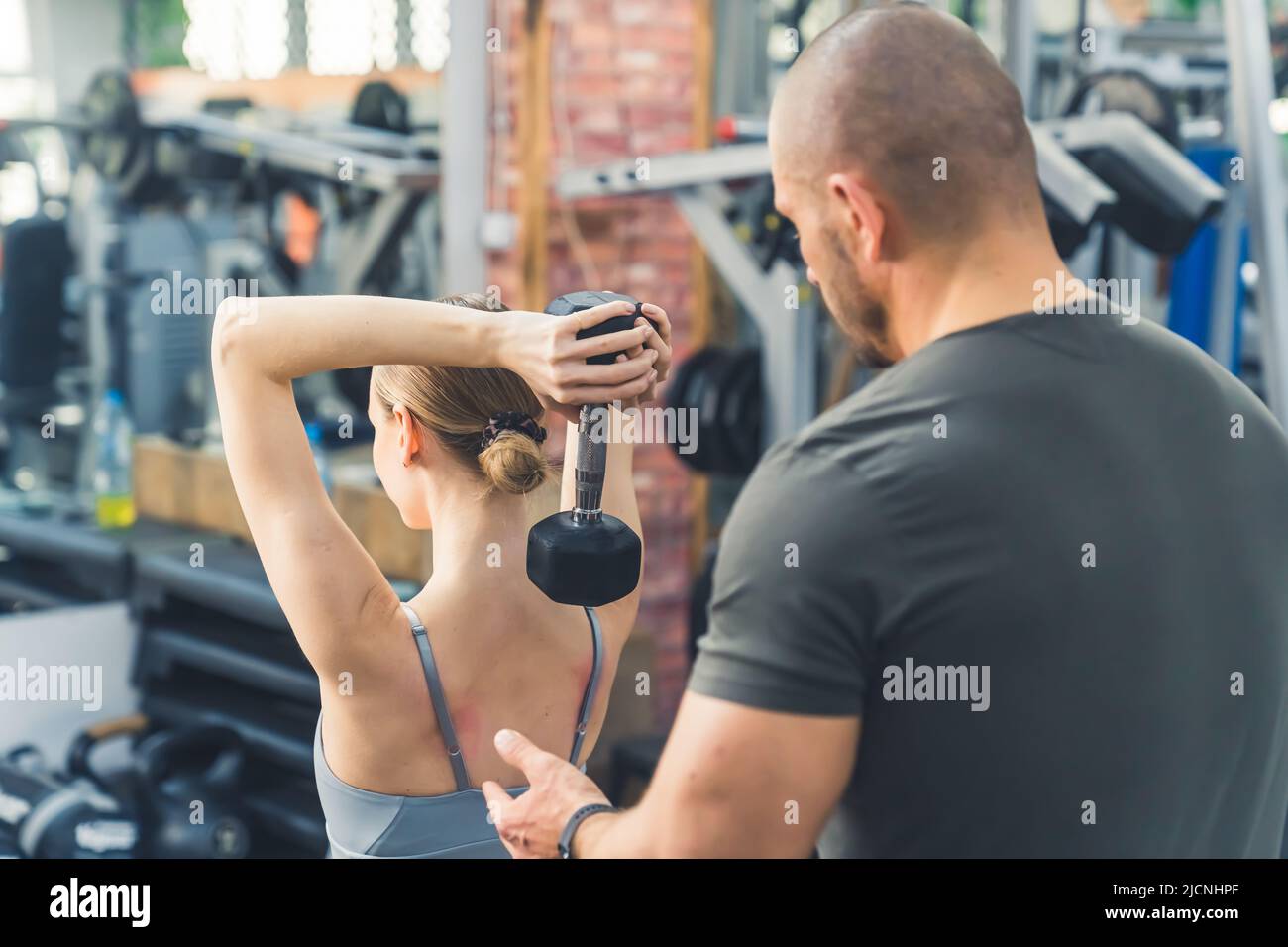 Indoor back shot in palestra di una giovane donna caucasica attraente sollevamento di un dumbbell, excercising triceps. Ragazzo muscolare calvo di mezza età che la sostiene. Foto di alta qualità Foto Stock