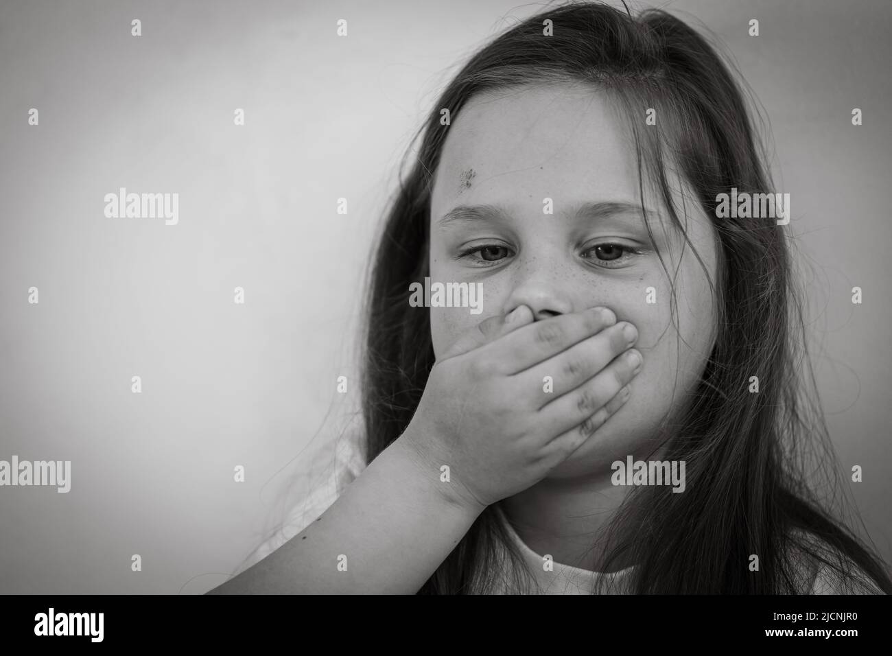 Ritratto bianco e nero di piccola ragazza triste livida con capelli lunghi e scuri che ricoprono la bocca con la mano. Protesta contro gli abusi. Foto Stock