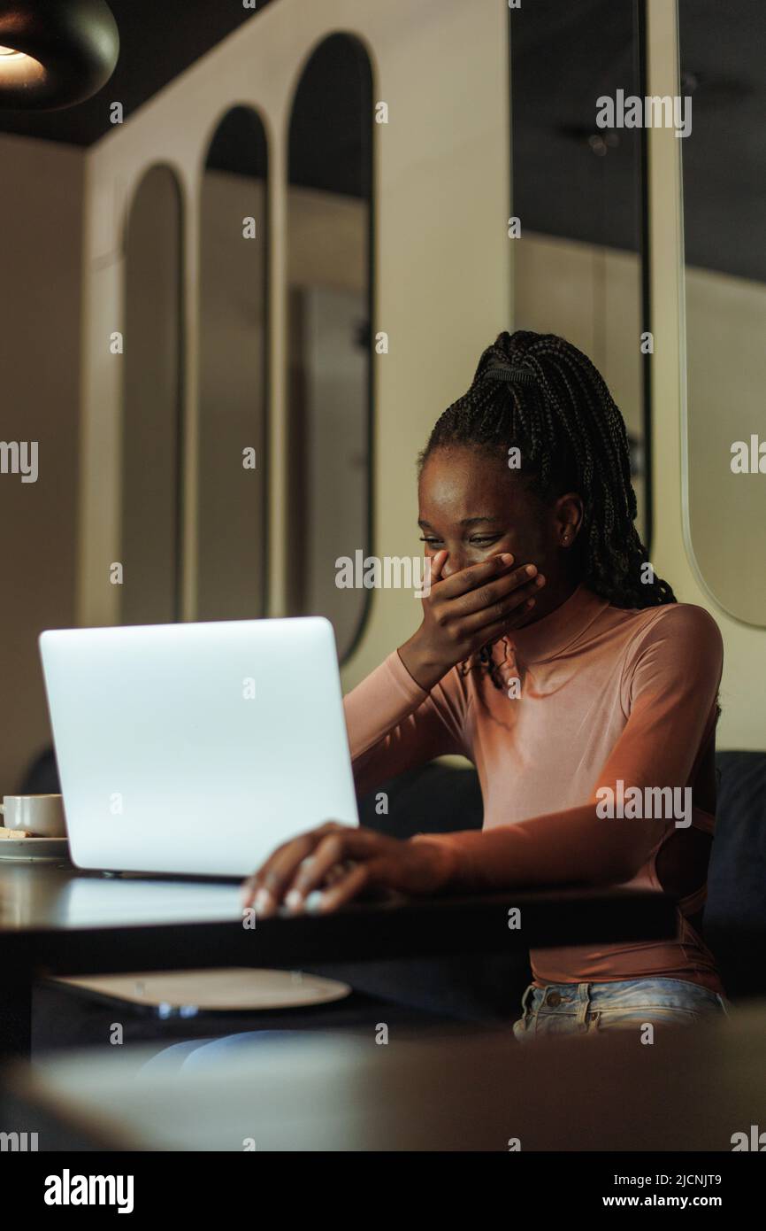 Ridendo millennial afro americano donna blogger, coprire la bocca con la mano, bere caffè, guardare il webinar sul portatile al caffè Foto Stock
