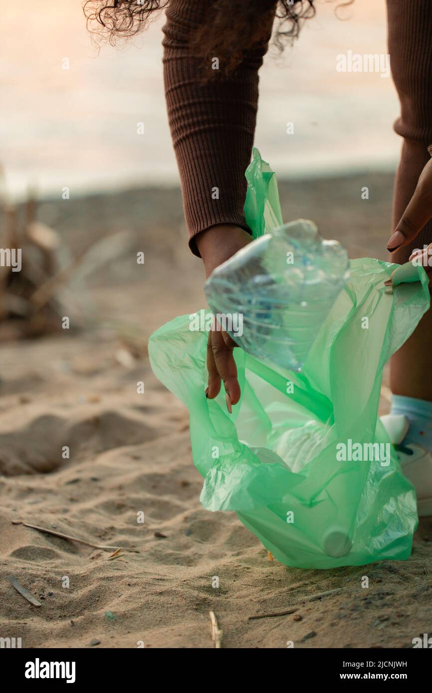 Donna africana irriconoscibile appoggiata giù per raccogliere i rifiuti dalla sabbia sulla spiaggia in sacchetto di plastica verde. Ecologia, inquinamento. Foto Stock