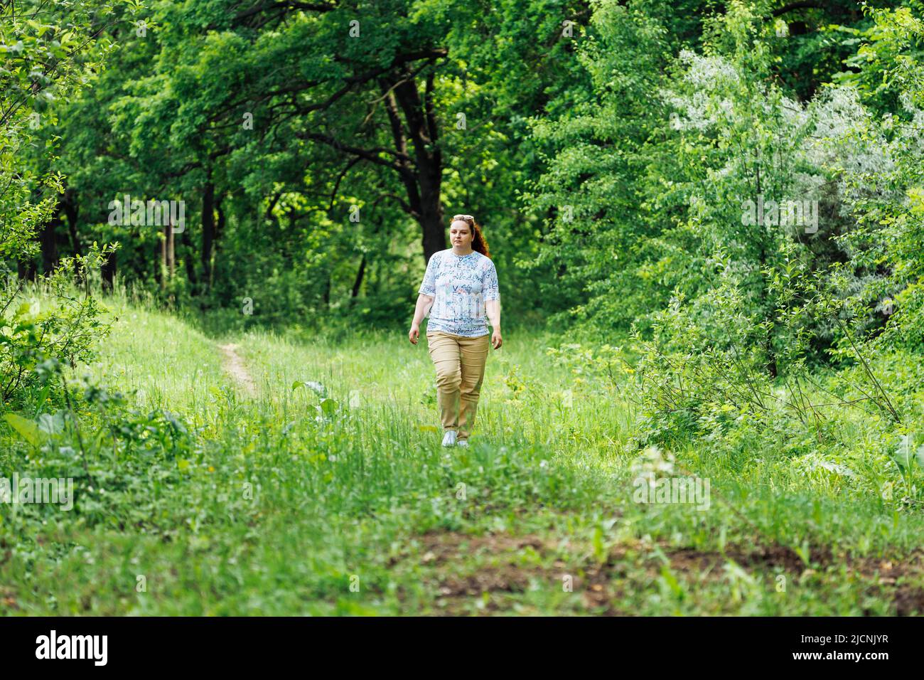 Ritratto di giovane donna buxom con lunghi capelli rossi ricci che camminano nella foresta del parco tra alberi verdi cespugli. Estate, natura. Foto Stock