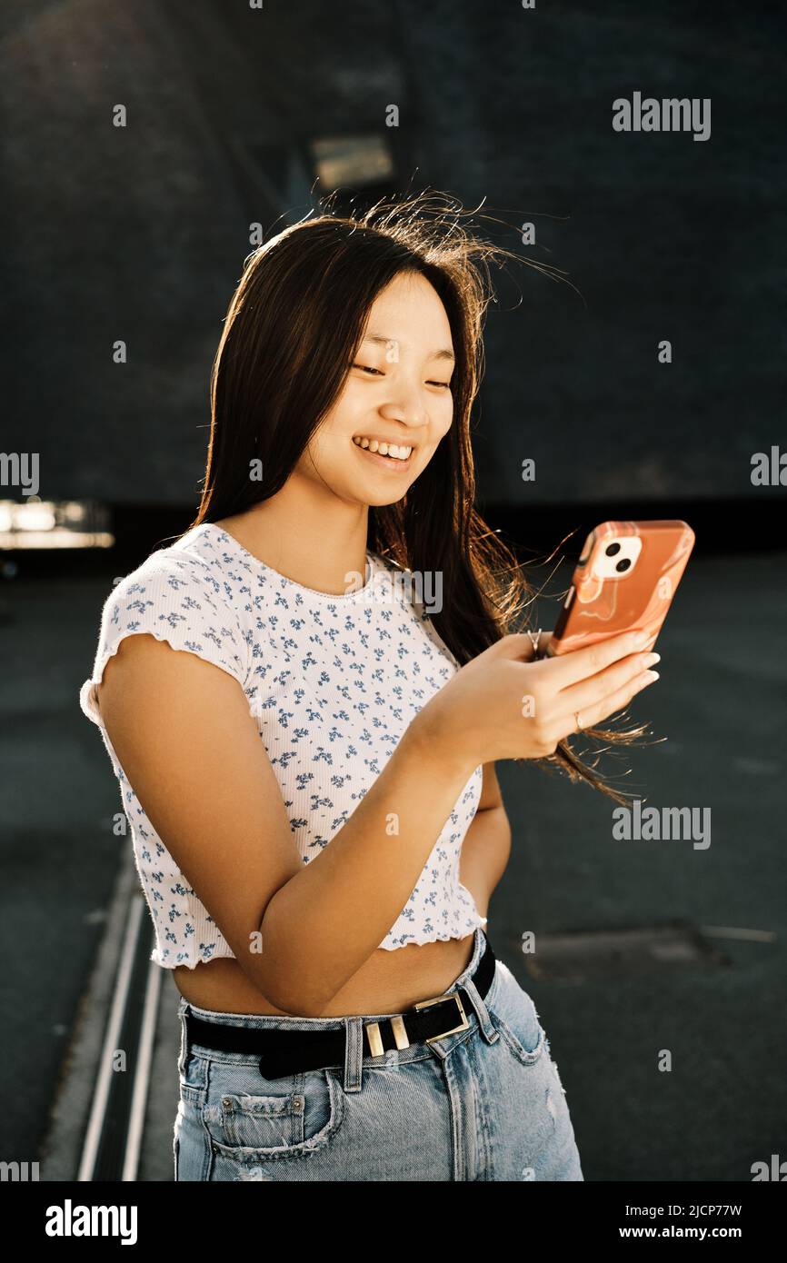 Giovane donna asiatica sorridente mentre usa il suo cellulare per strada. Foto Stock
