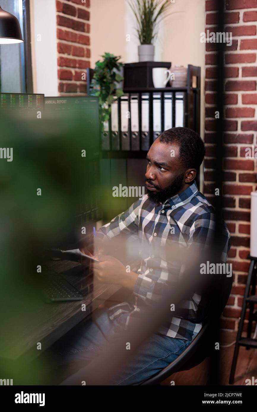 Operatore di hedge fund che analizza la tendenza del mercato mentre si siede alla stazione di lavoro avendo appunti di documentazione. Trading Analyist monitora i dati finanziari in tempo reale per prevedere il prossimo crollo dei prezzi. Foto Stock