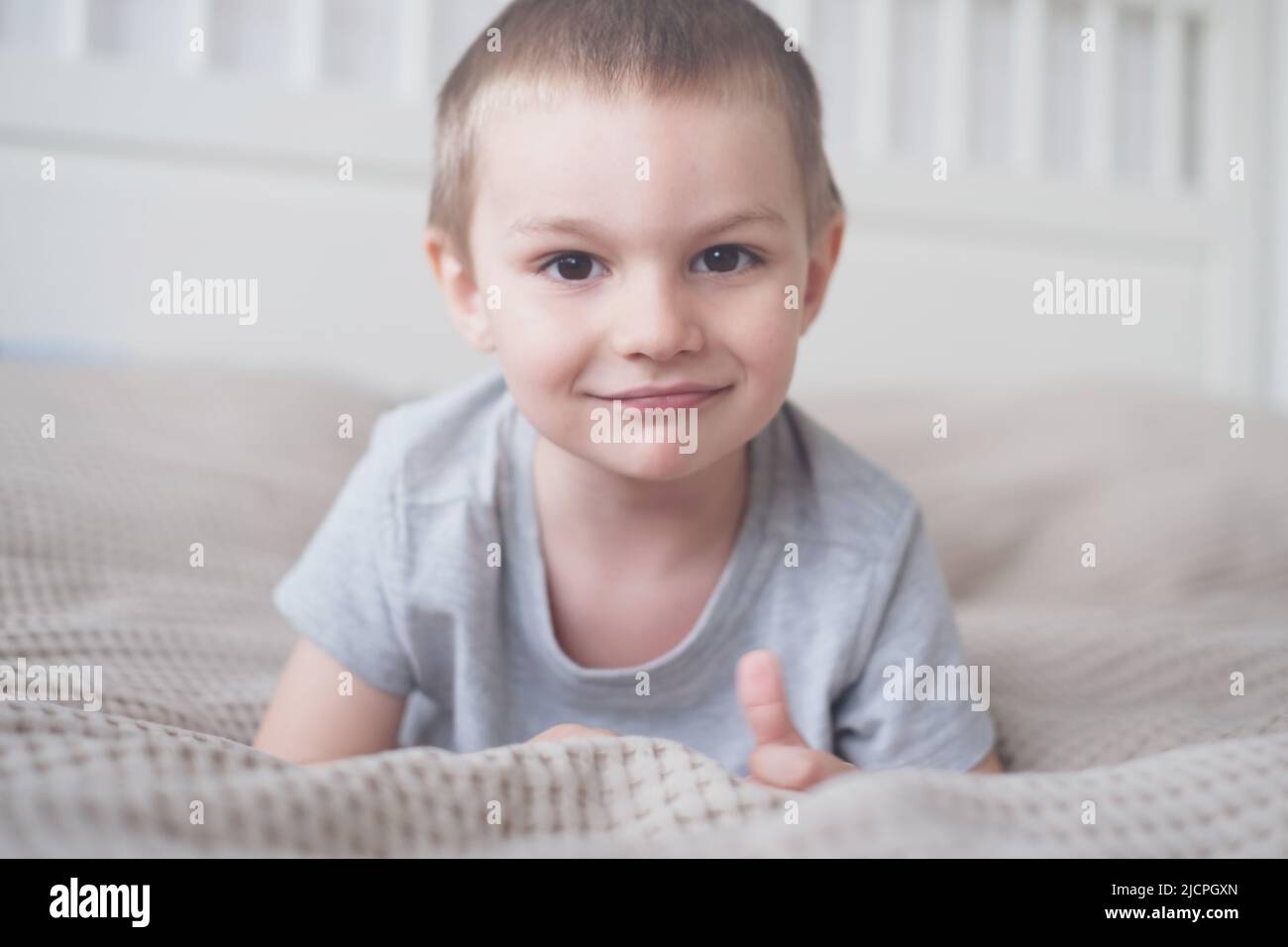 Primo piano di un piccolo ragazzo caucasico felice in una t-shirt grigia si trova sul letto e sorride Foto Stock