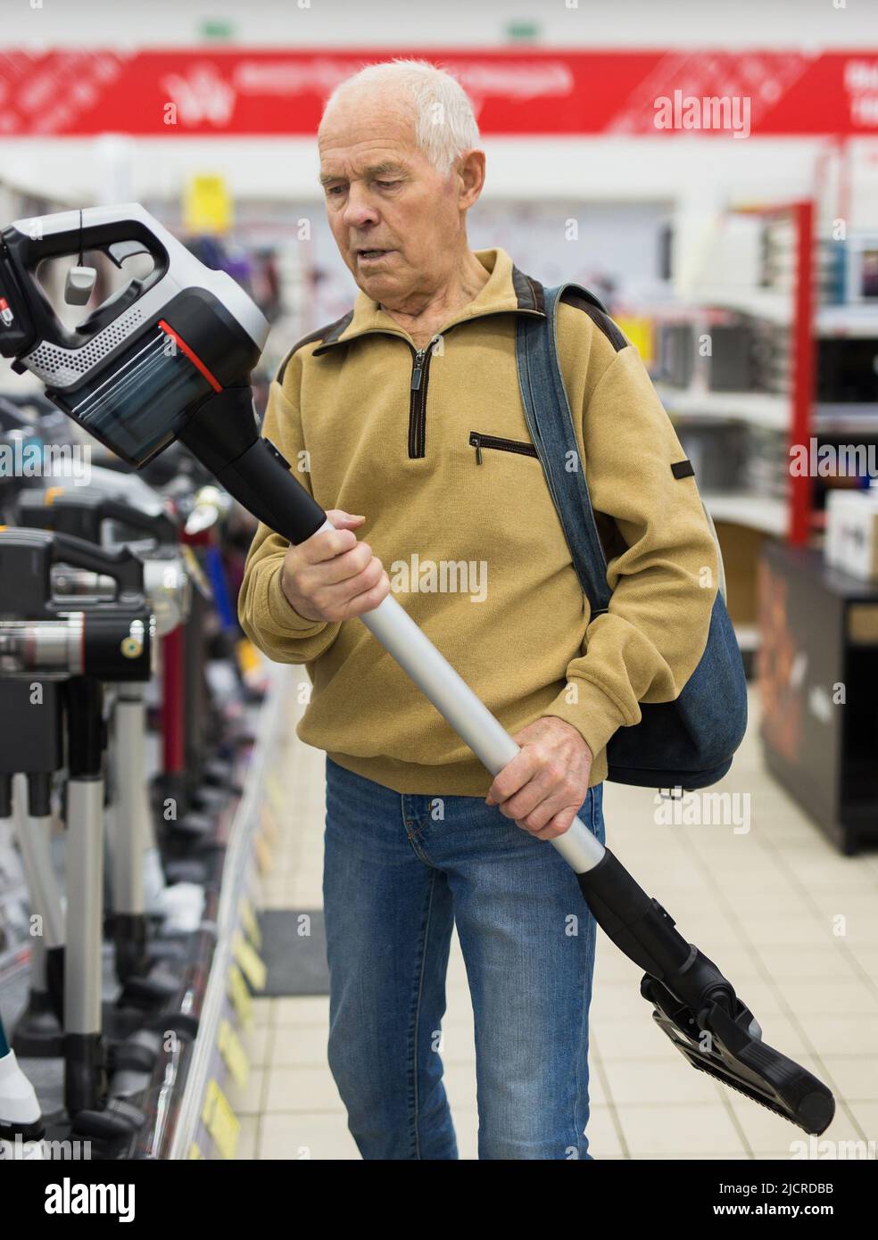 Uomo anziano in grigio pensionato guardando verticale aspirapolvere hoover al banco in showroom di elettrodomestici ipermercato reparto Foto Stock