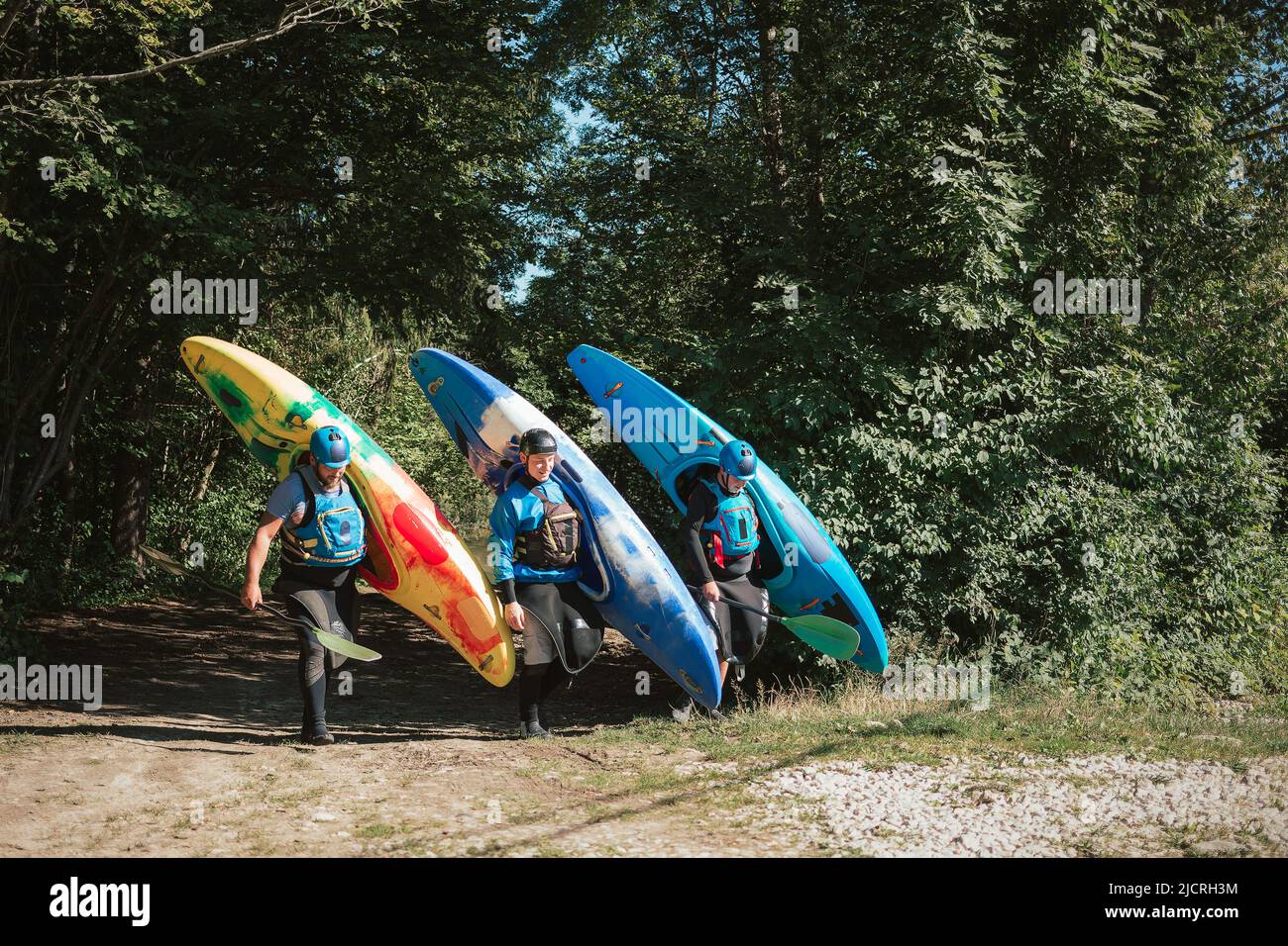 Tre kayak da diporto con caschi e giubbotti salvagente si muovono dal fiume attraverso il bosco verde, trasportando kayak e pagaie Foto Stock