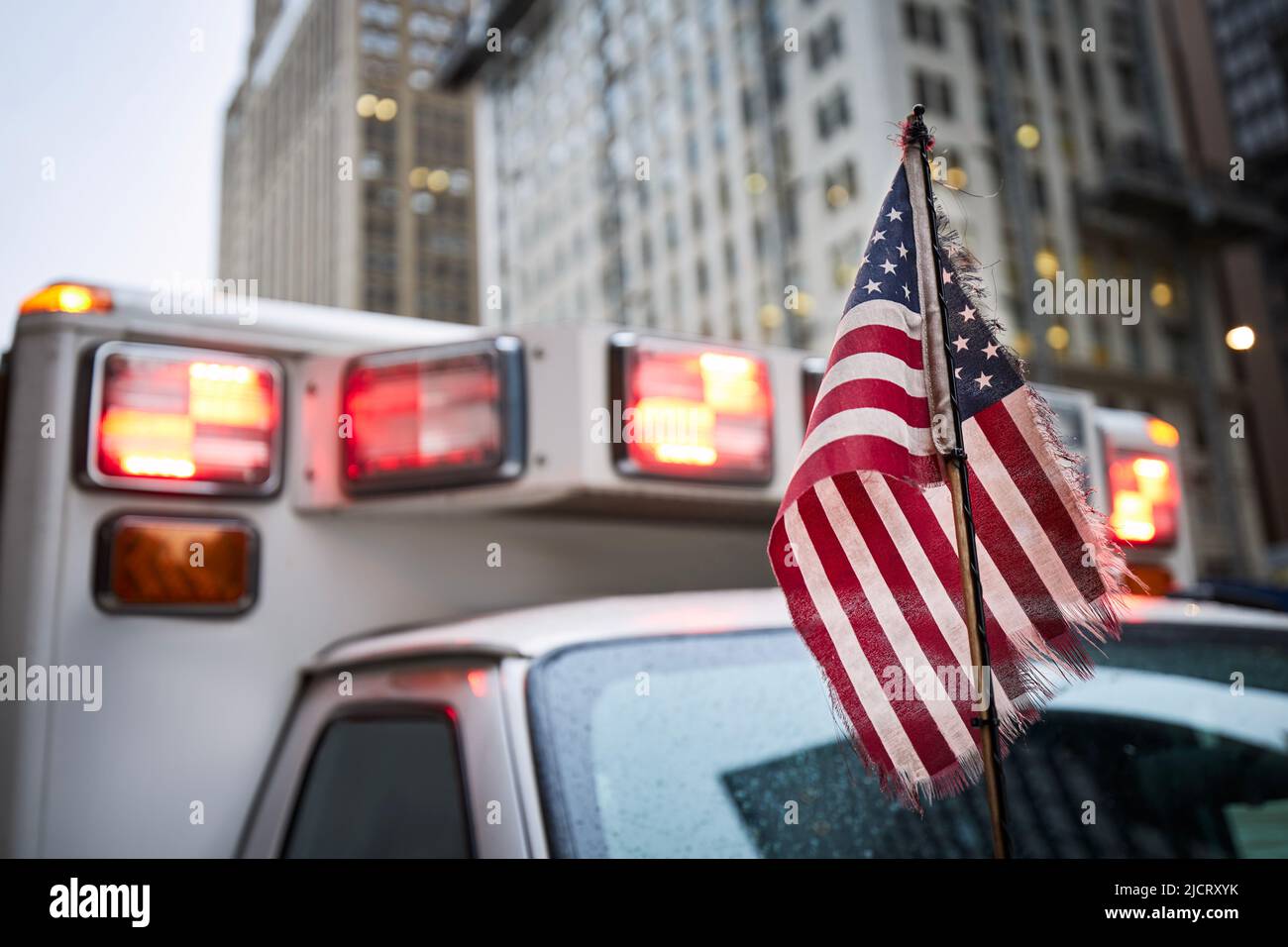 Primo piano della bandiera americana contro la luce lampeggiante rossa della sirena dell'auto dell'ambulanza. Temi di emergenza, soccorso e aiuto. Foto Stock
