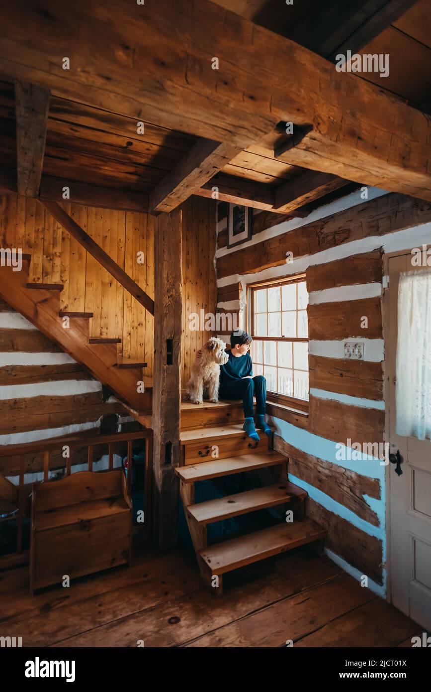 Ragazzo e cane seduti sulle scale di casa di legno cabina guardando fuori finestra. Foto Stock