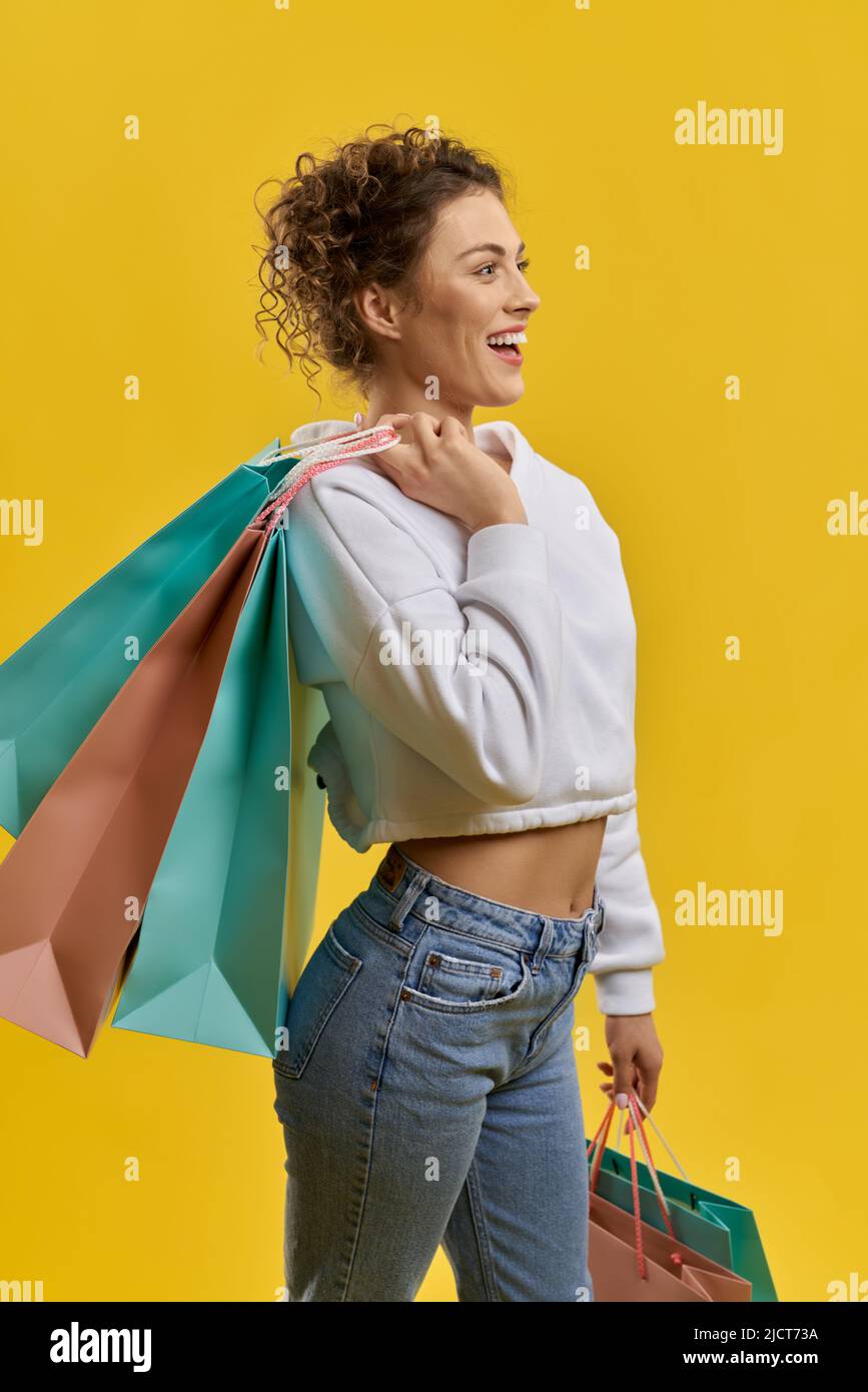 Donna curly sicura che porta borse shopping a spalla, sorridente al coperto. Vista laterale di una donna sottile shopper che guarda felice, isolato su sfondo arancione studio, con spazio per la copia. Concetto di shopping. Foto Stock