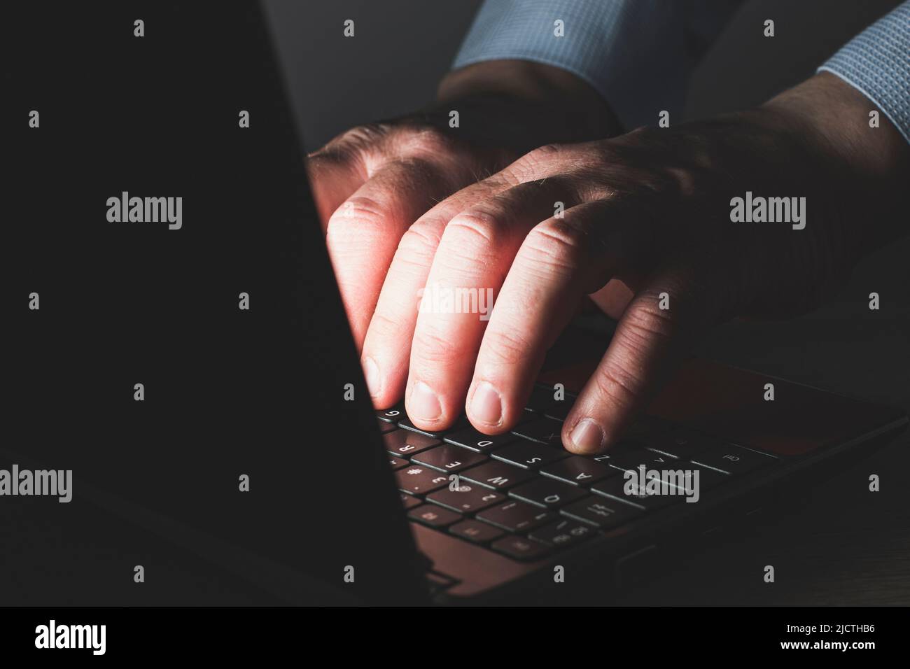 Le mani di un uomo caucasico irriconoscibile in una maglietta blu che digita su una tastiera di un computer portatile. La scena è scura e le mani sono illuminate dalla luce Foto Stock