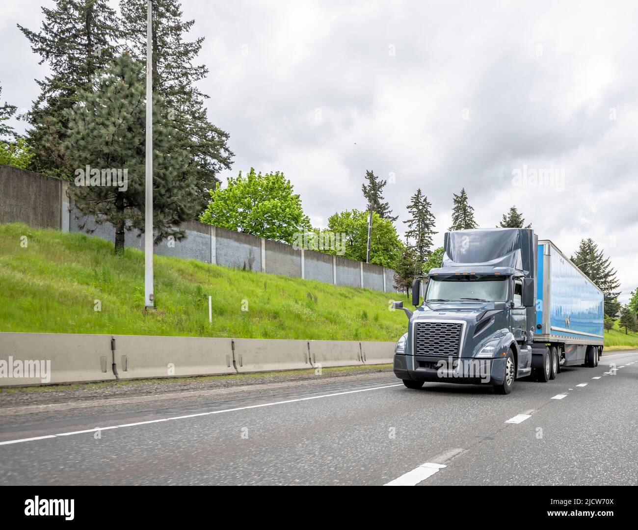 Trattore semi-camion grigio industriale con carro grande classico e carico per il trasporto di carichi commerciali in semirimorchio con furgone a secco che gira sull'ampia strada rettilinea r Foto Stock