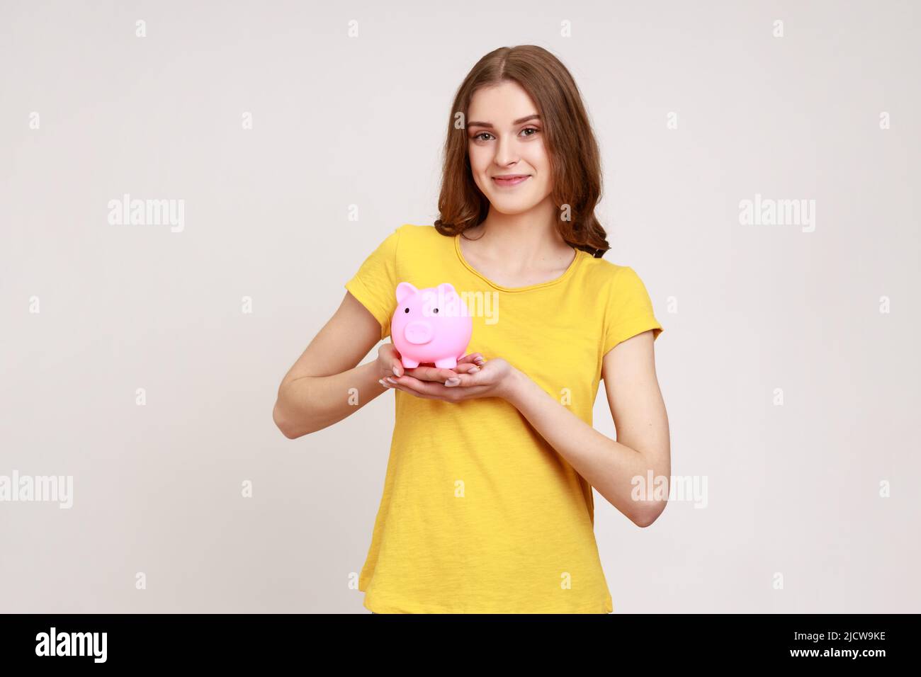 Ragazza sorridente attraente con i capelli marroni che tengono la banca del piggy, essendo felice con i risparmi di soldi, indossando la T-shirt di stile casual gialla. Studio interno girato isolato su sfondo grigio. Foto Stock