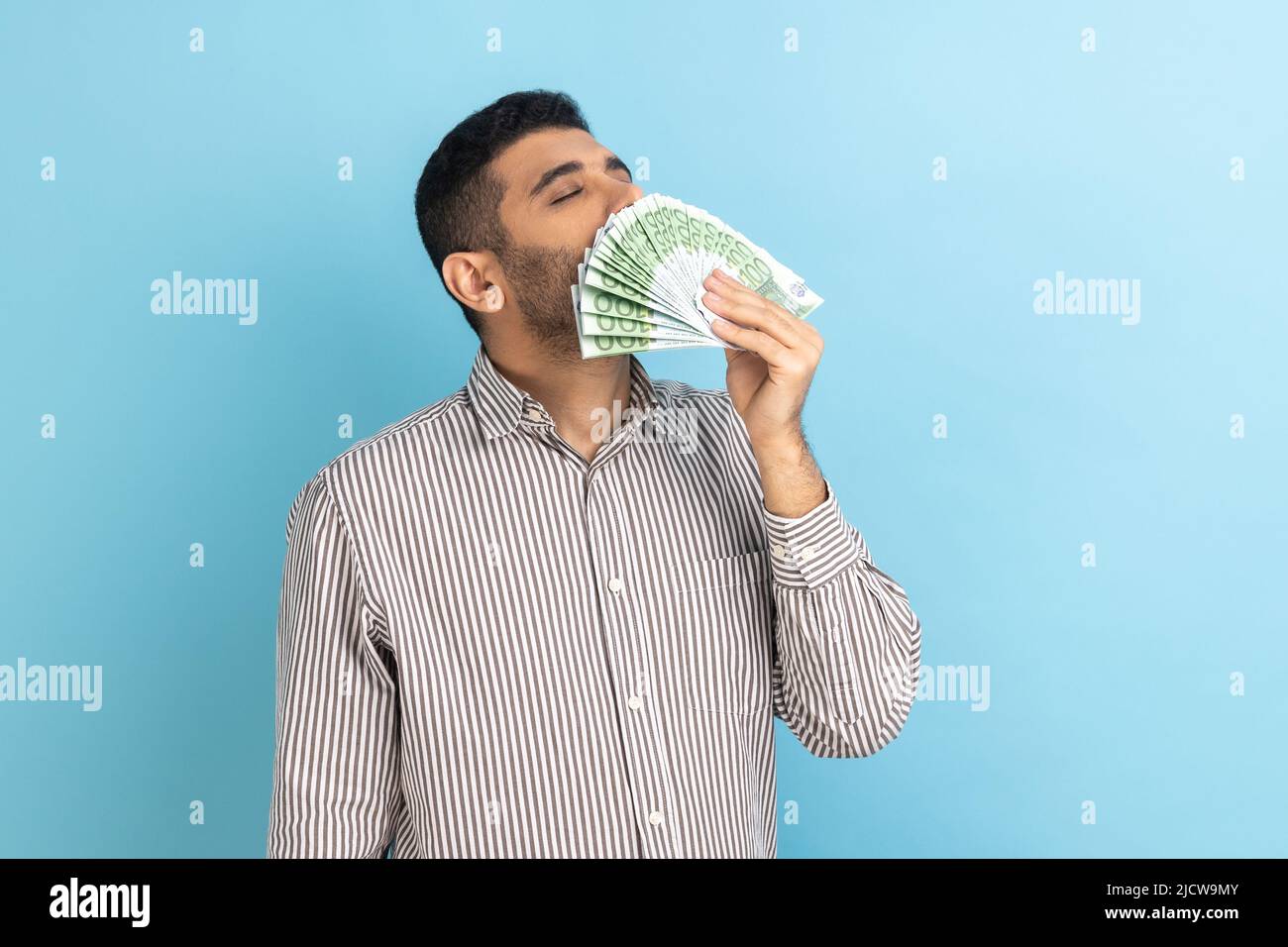 Ritratto di avarioso uomo d'affari che puzzava banconote in euro, godendo della vittoria della lotteria, grande profitto e vita ricca, indossando una camicia a righe. Studio interno girato isolato su sfondo blu. Foto Stock