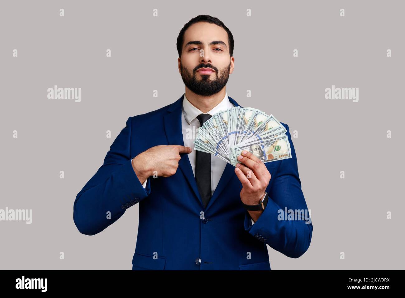 Ritratto di ricco auto fiducioso ported uomo d'affari che tiene le fatture dei dollari e che punta a se stesso con orgoglio, vestito ufficiale di stile. Studio interno girato isolato su sfondo grigio. Foto Stock