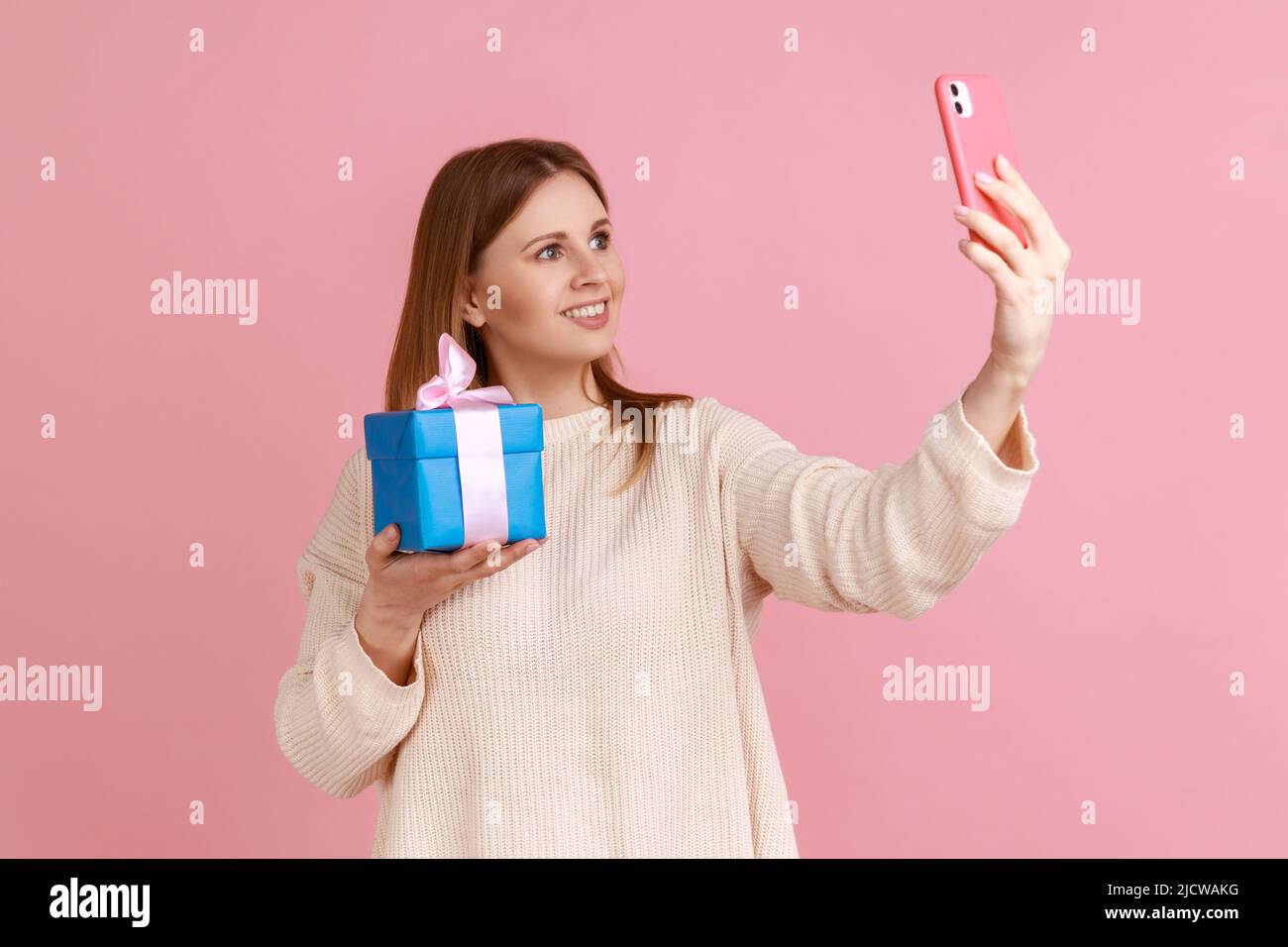 Donna sorridente attraente che prende selfie con il suo regalo in scatola blu o che vanta il suo regalo con i seguaci mentre ha livestream, indossando il pullover bianco. Studio interno girato isolato su sfondo rosa. Foto Stock