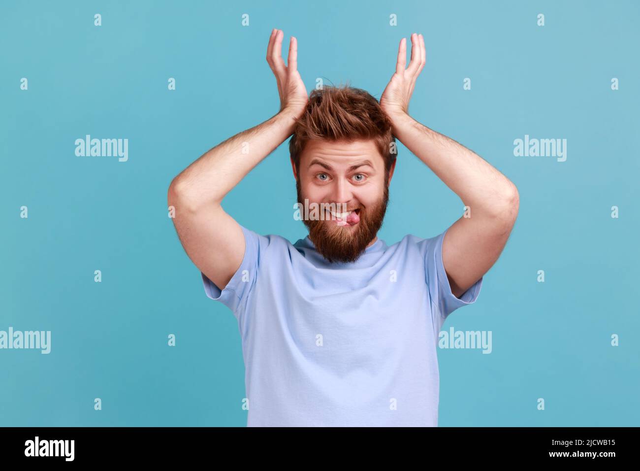 Ritratto di uomo positivo barbuto in piedi con corna di corna sopra la testa e mostrando lingua fuori, guardando con comica espressione umoristica. Studio interno girato isolato su sfondo blu. Foto Stock