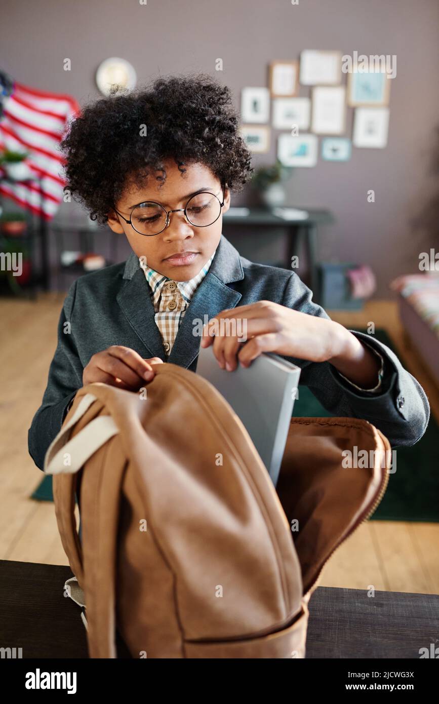 Scolari afroamericani in occhiali che imballano i libri nel suo zaino al tavolo nella stanza prima di andare a scuola Foto Stock