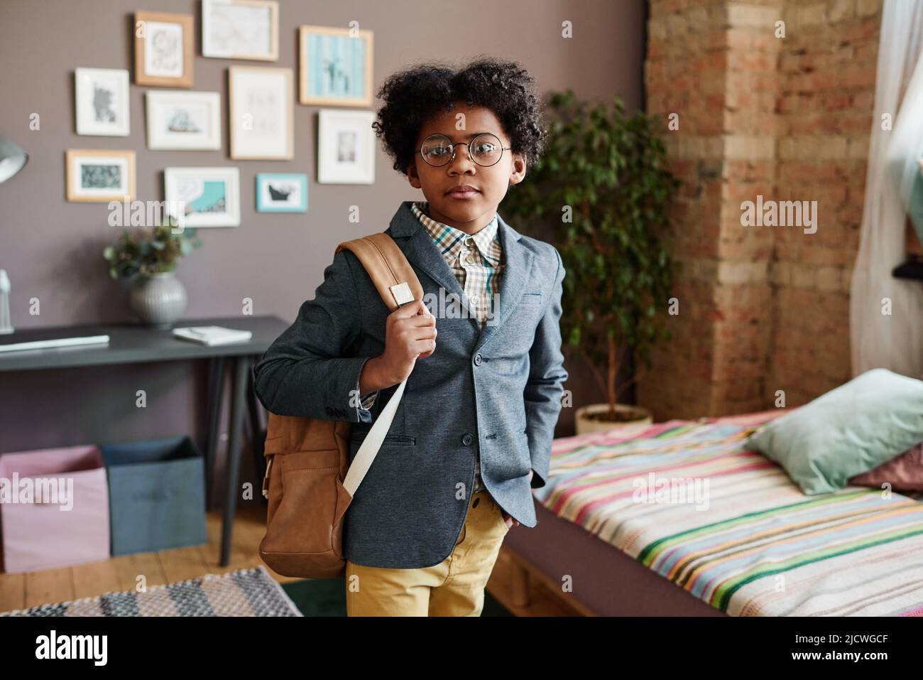 Ritratto di scolaro africano in tuta con borsa di studio guardando la macchina fotografica mentre si trova in piedi nella stanza, lui va a scuola nella sua prima volta Foto Stock