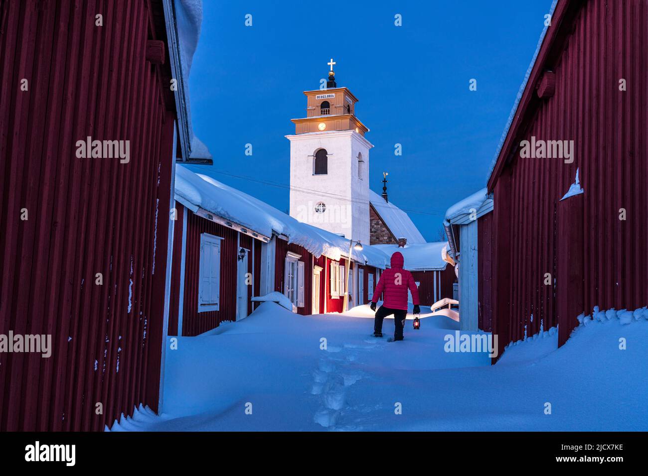 Persona che ammira il campanile in piedi nella neve profonda tra cottage rossi al tramonto, Gammelstad Church Town, sito patrimonio dell'umanità dell'UNESCO, Lulea, Svezia Foto Stock