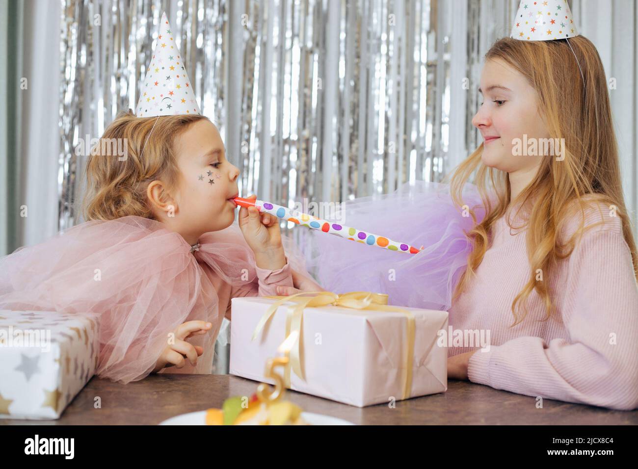 Due simpatiche sorelle celebrano il compleanno del più giovane, che ha compiuto 5 anni. Ragazze che si divertono con il corno festivo, ci sono scatole regalo e torta di fronte Foto Stock