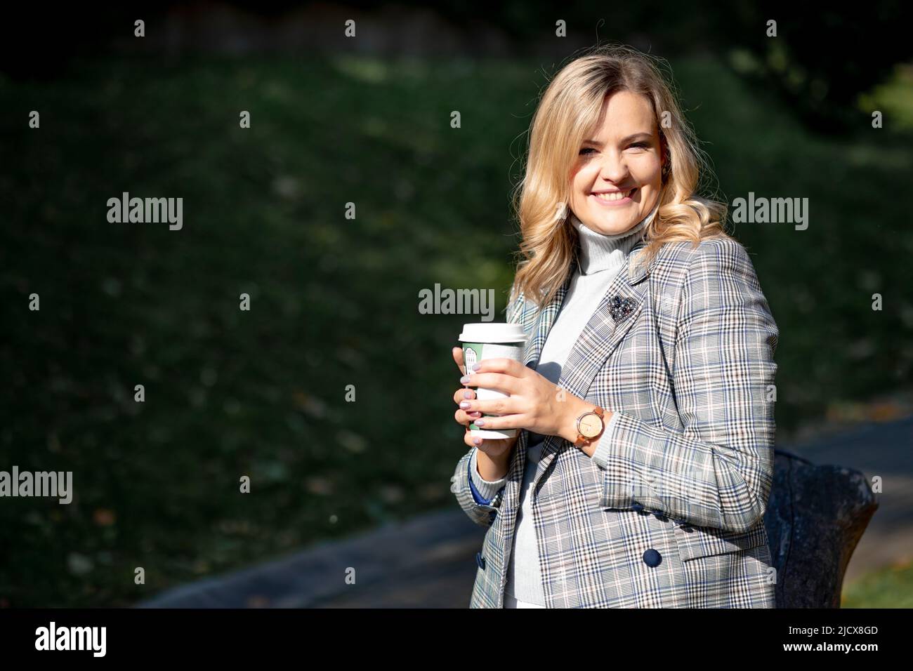 La calda luce del sole illumina il volto felice della bionda in una giacca grigia tweed e un pullover in cashmere abbinato, tenendo in mano una tazza con una bevanda calda. Femminilità Foto Stock