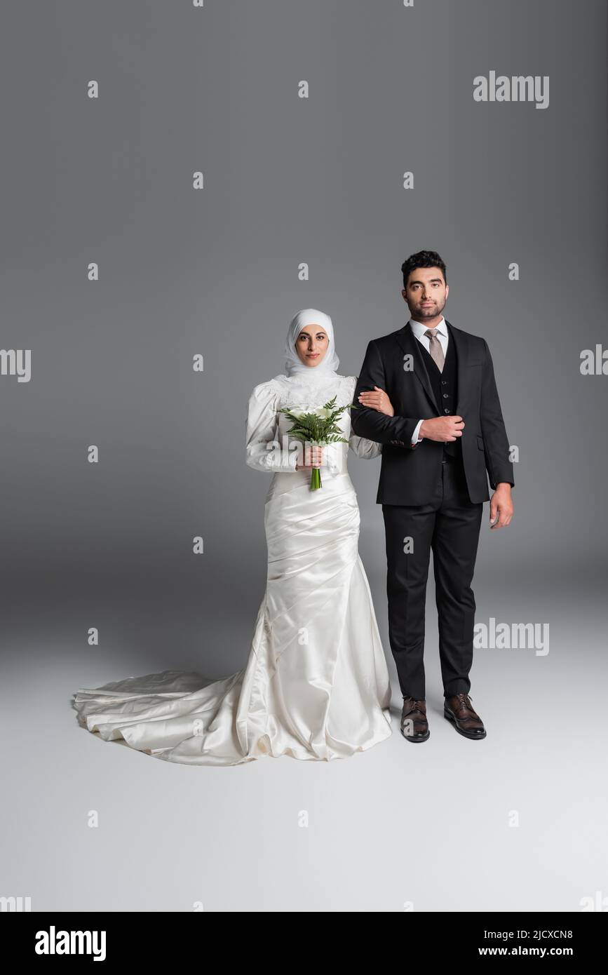 l'intera lunghezza dello sposo in vestito in piedi con la sposa musulmana con il bouquet di nozze dei fiori del giglio del calla sul grigio Foto Stock