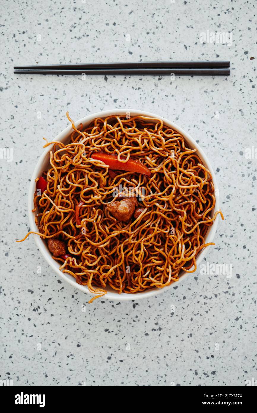 vista ad angolo alto di un paio di bacchette nere e un piatto biancastro con alcuni spaghetti di pollo yakisoba su una superficie bianca in pietra chiazzata Foto Stock