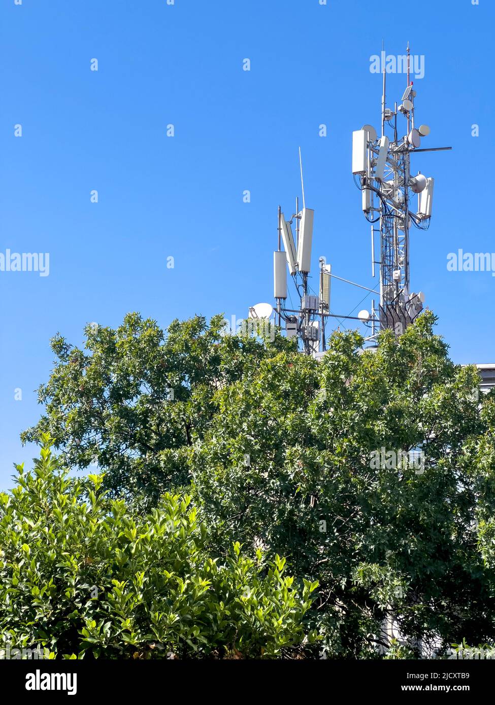 Antenna GSM Tower, spazio copia. Tecnologia delle telecomunicazioni per 5G, 4G, 3G. Telefono cellulare, antenne TV, tetto dietro l'albero verde. Cielo azzurro soleggiato Foto Stock