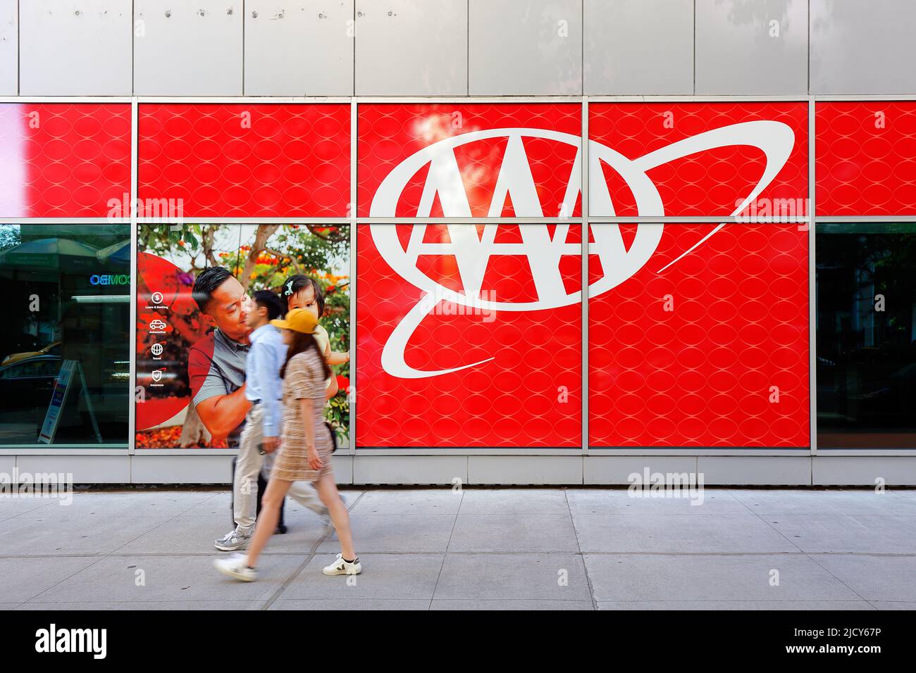Una coppia asiatica cammina oltre un grande logo AAA fuori dall'agenzia di assicurazione American Automobile Association, 1881 Broadway, New York, NY. Foto Stock