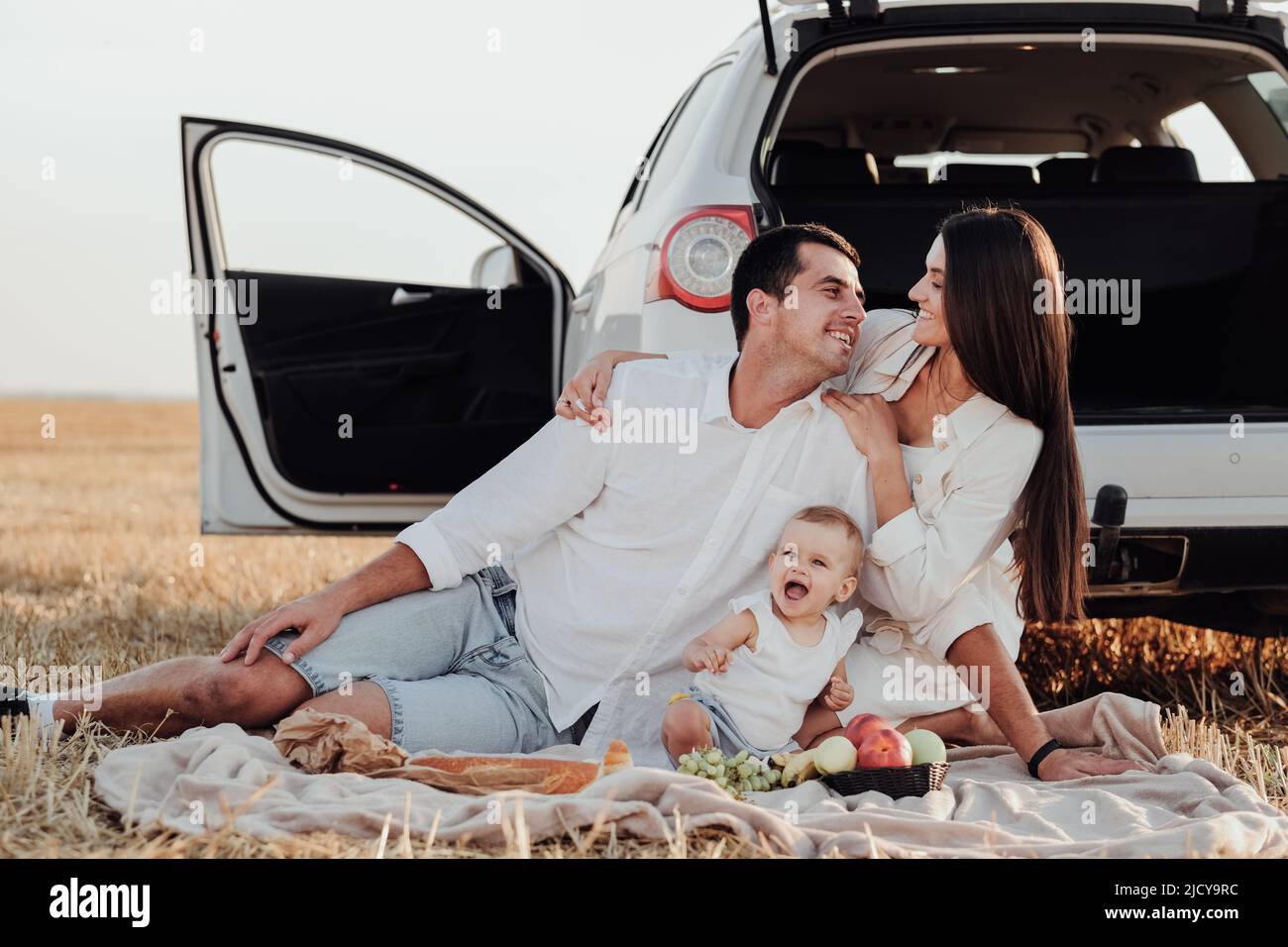 Sorridendo giovane famiglia con bambino bambino che ha un picnic fuori della città, mamma e papà con la loro figlia vestita simile seduto vicino alla macchina all'aperto Foto Stock