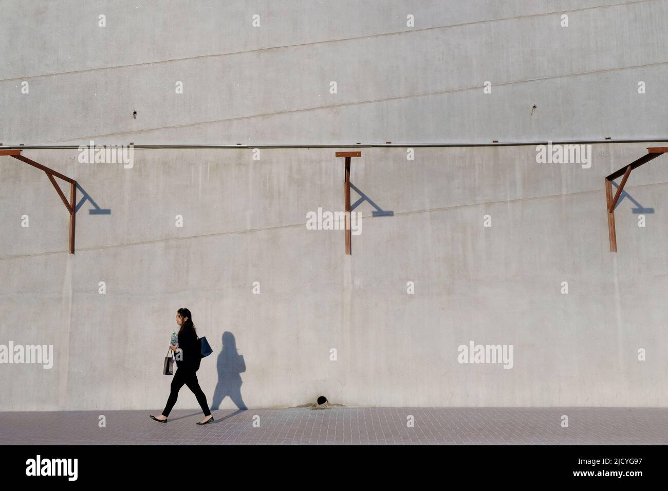 Donna che cammina sul marciapiede, accanto alla costruzione con muro di cemento bianco con barre di metallo per contenere grandi annunci e cartelloni. Spazio di copia orizzontale. Foto Stock