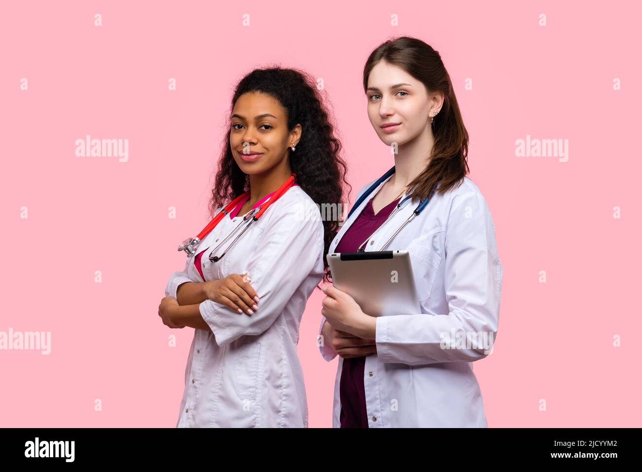 Ritratto due metà-girato amichevole medico GP femmina con le braccia incrociate su torace e tablet digitale su rosa Studio Wall. Diverse etnie Medical Tea Foto Stock