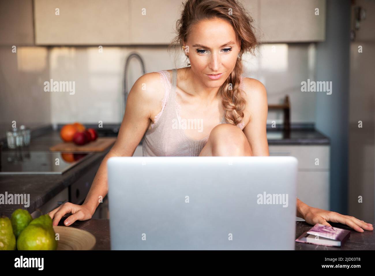 Una giovane bella donna di buon umore comunica via video link su un computer portatile in cucina della casa in prima serata al tramonto. Foto Stock
