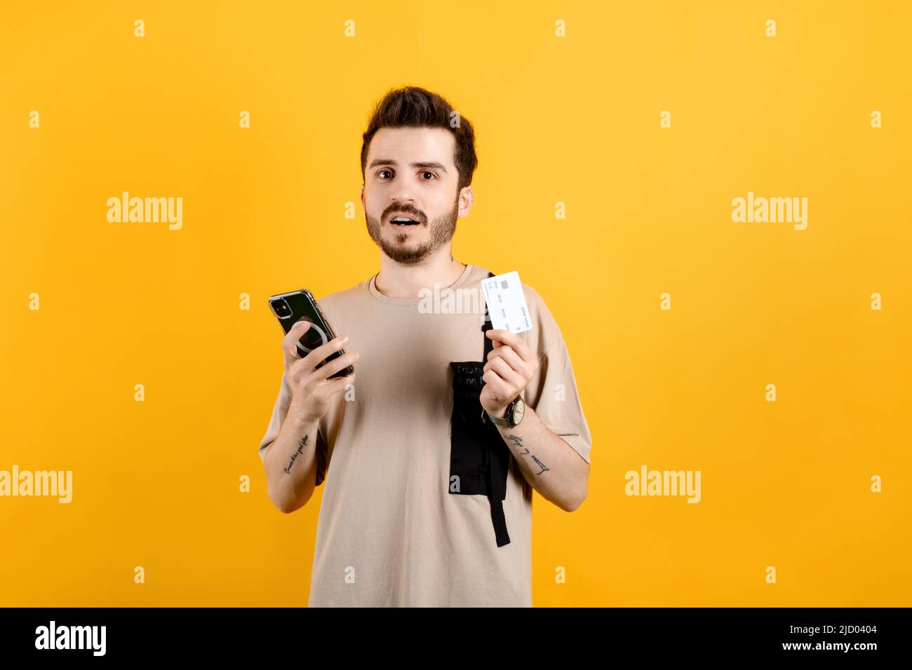 Uomo caucasico che indossa tee beige posa isolato su sfondo giallo tenendo carta di credito e smartphone con volto deluso, shrugging, fallimentare Foto Stock