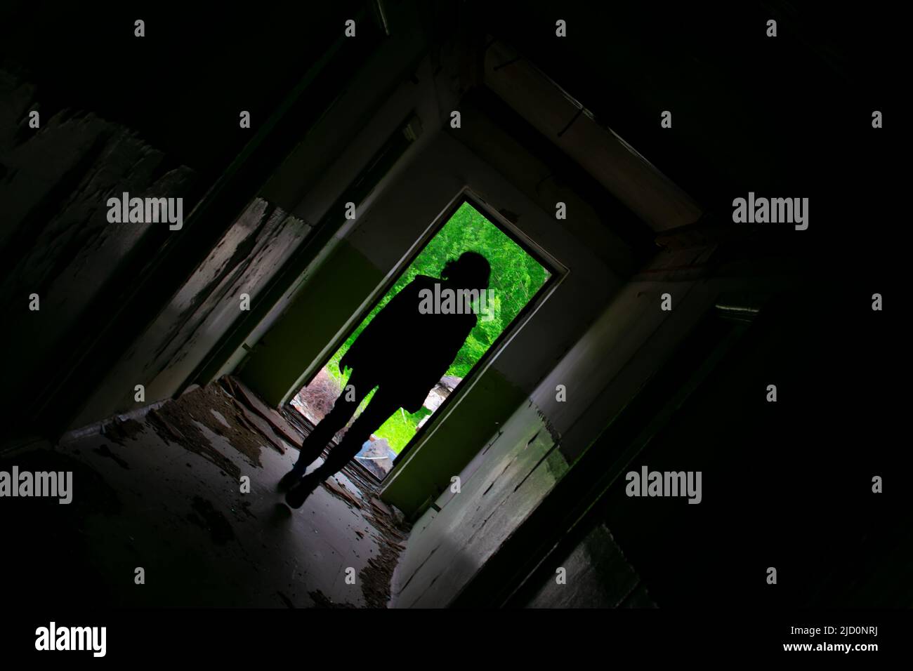 una vista di un corridoio scuro in pendenza di un edificio abbandonato con una silhouette di una ragazza che cammina fino all'uscita, in cui il verde luminoso fogliame di alberi Foto Stock
