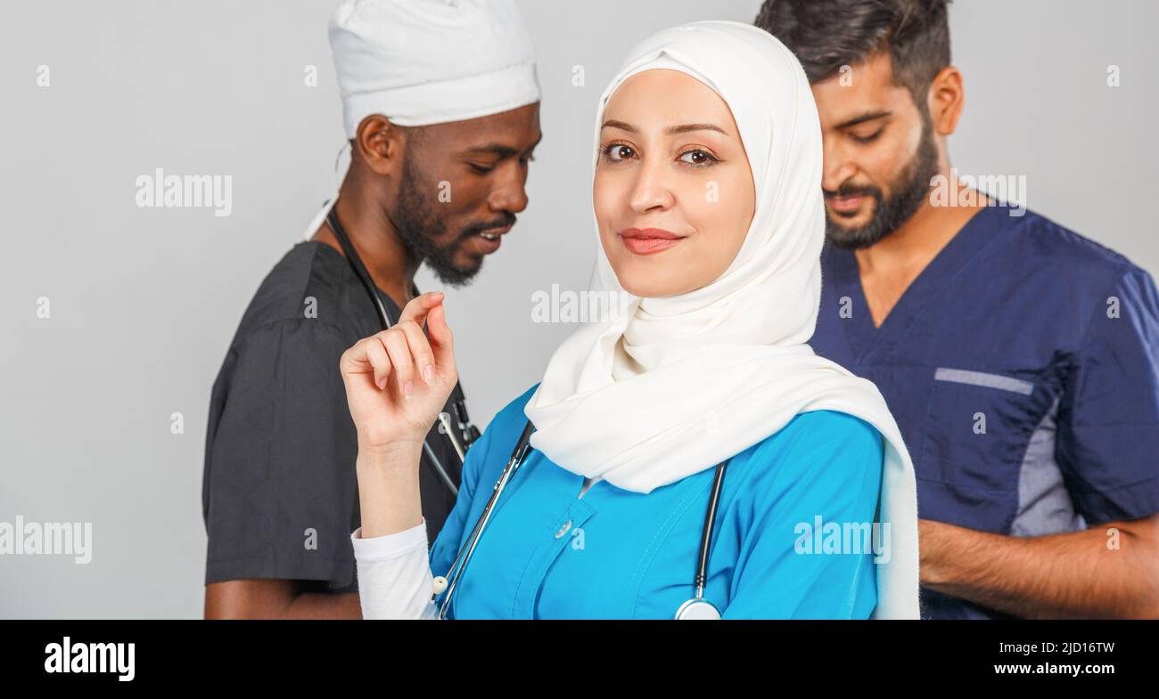 Gruppo di paramedici sorridenti su sfondo grigio. donna medico musulmana con colleghi chirurgo africano e indiano Foto Stock