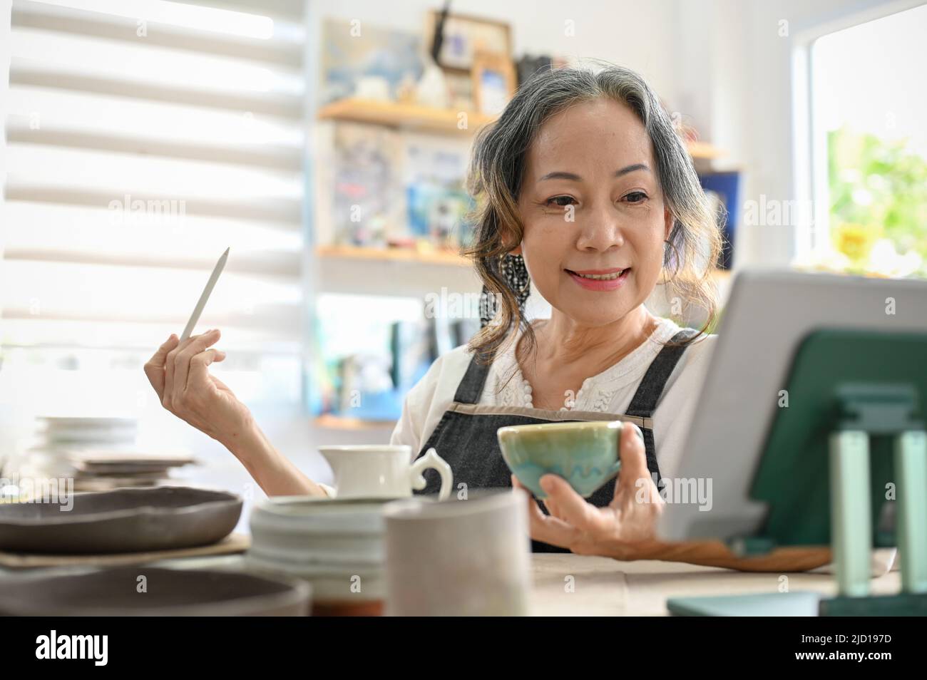 Bella donna asiatica invecchiata imparare una classe di pittura di ceramica on-line via tablet e dipingere la sua tazza di ceramica nel suo studio di casa. Attività creative Foto Stock