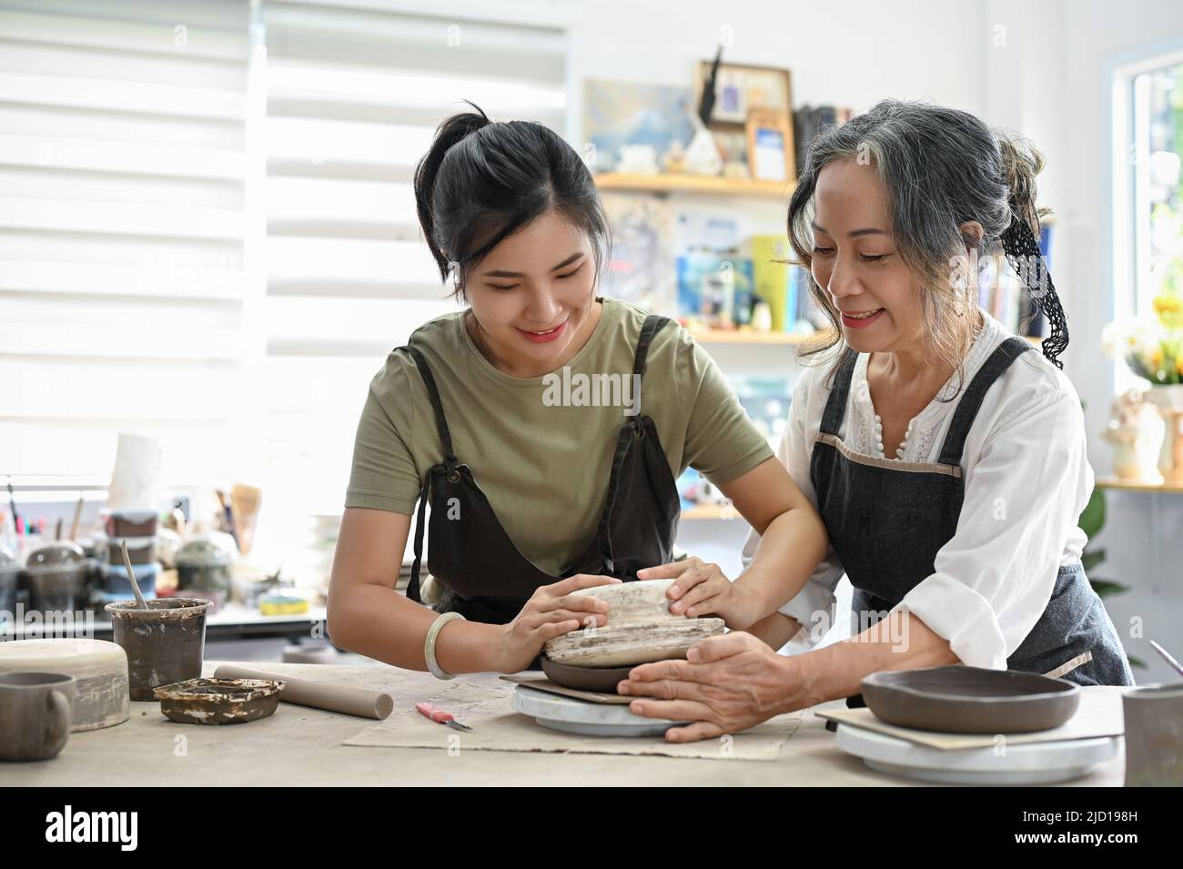 Insegnante professionale di età femminile che insegna una donna piuttosto giovane a fare una ciotola di ceramica con argilla nel laboratorio. Attività creative e hobby conce Foto Stock