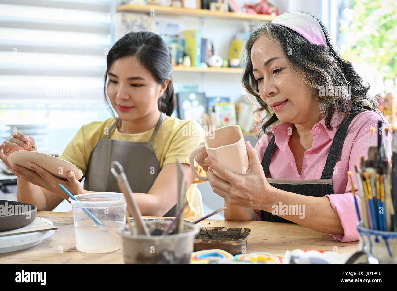 Bella giovane donna asiatica che dipinge una lastra di ceramica di colore acrilico con sua nonna in officina. Buona famiglia con attività creative c Foto Stock