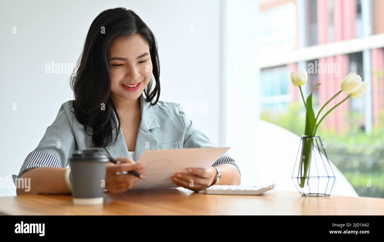 Affascinante giovane donna d'affari asiatica che rivede un rapporto di proposta di affari, analizzando il documento finanziario nell'ufficio moderno. Foto Stock