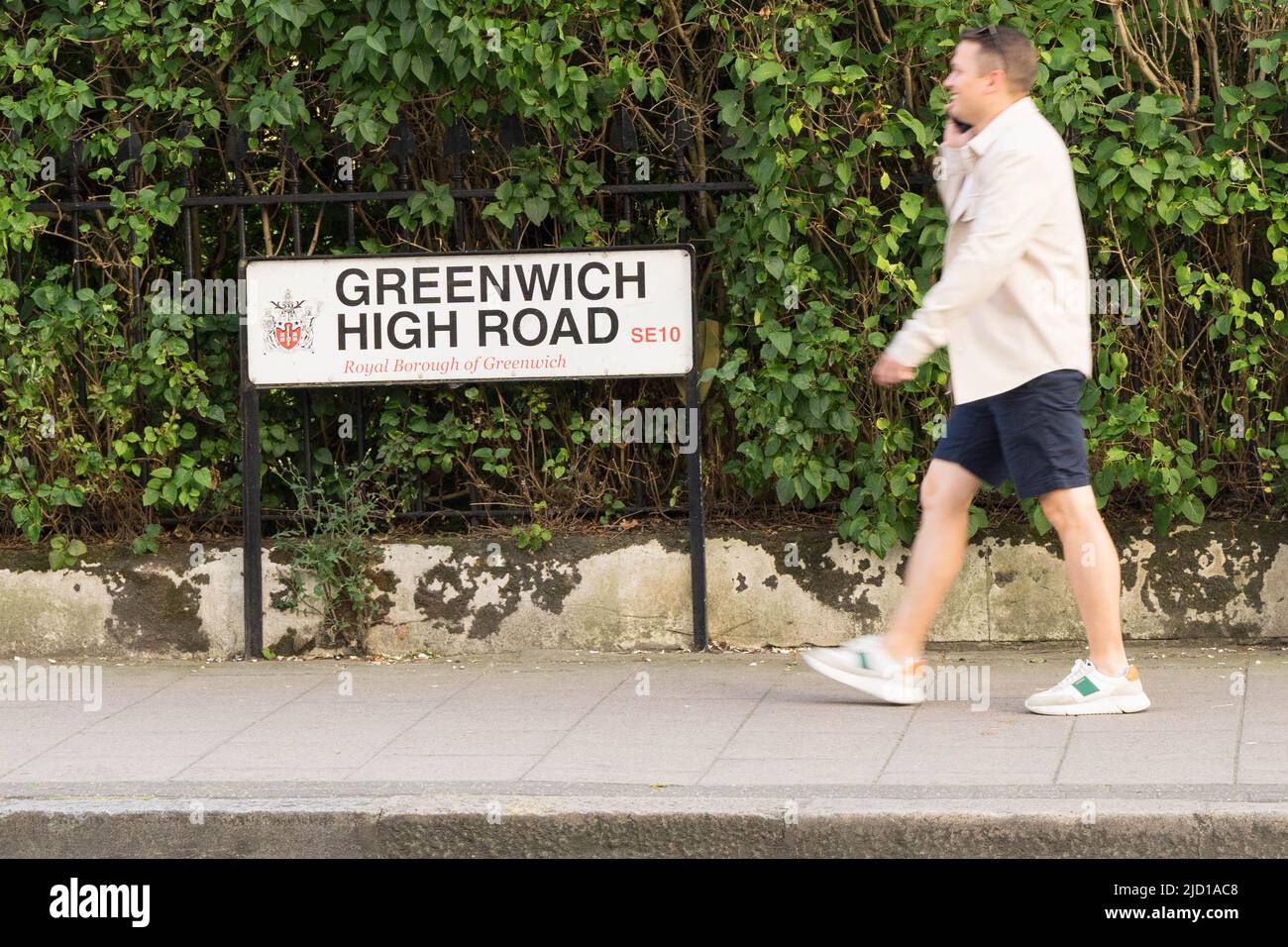 Un uomo in blu scuro corto e un paio di allenatori cammina sul marciapiede con un cartello stradale per Greenwich High Road London Inghilterra Foto Stock