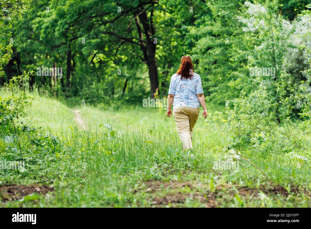 Ritratto di giovane donna buxom con lunghi capelli rossi ricci che camminano nella foresta del parco tra alberi verdi cespugli. Estate, natura. Foto Stock