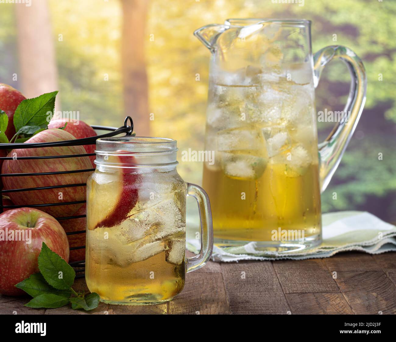 Bicchiere di succo di mela con fetta di mela e ghiaccio e un cestino di mele e caraffa su un tavolo di legno con sfondo rurale estivo Foto Stock