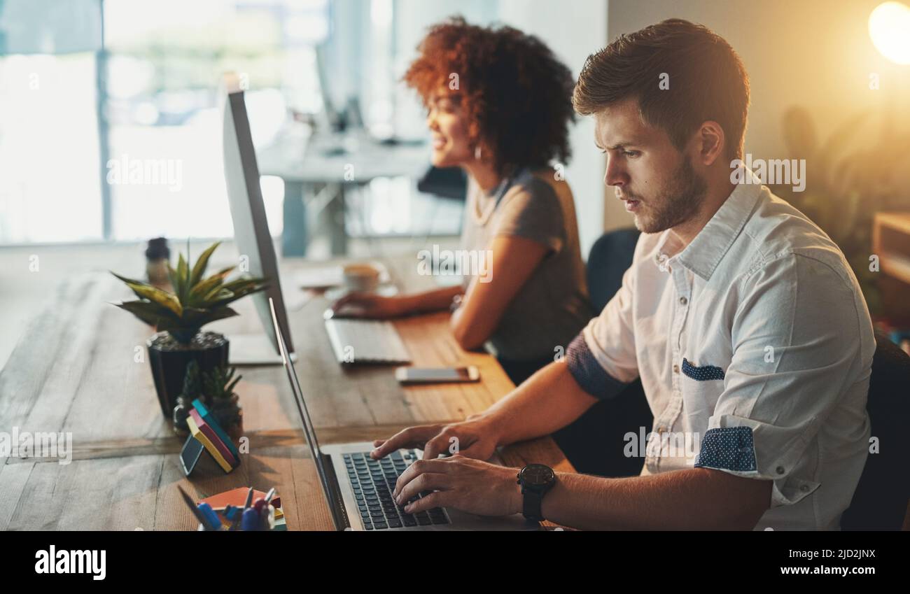 Mettendo nelle ore. Scatto di un giovane designer che lavora in ritardo con il suo collega sfocato sullo sfondo. Foto Stock